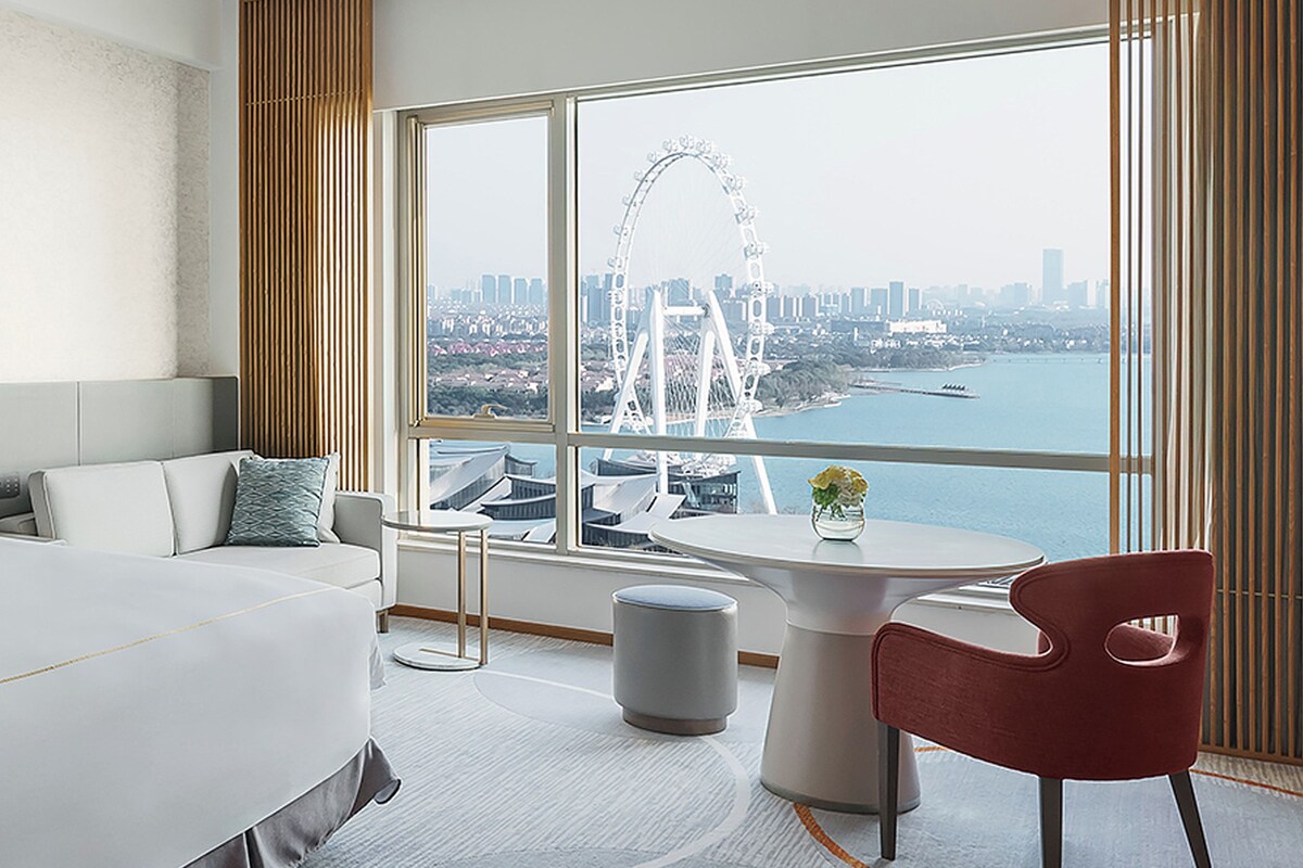 Habitación de hotel con una cama grande, un sofá, una mesita con un jarrón y vistas a una noria y al paisaje urbano.
