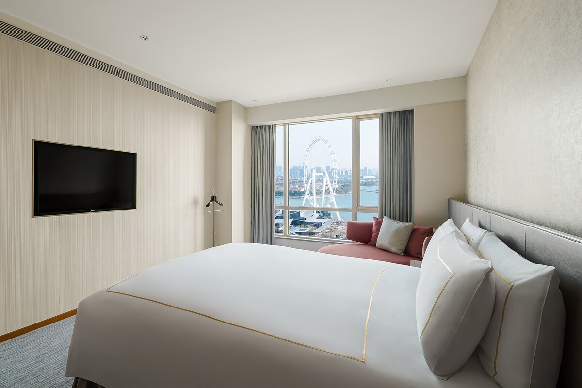 A hotel room with a large bed, a flat-screen TV mounted on the wall, and a view of a Ferris wheel through the window.