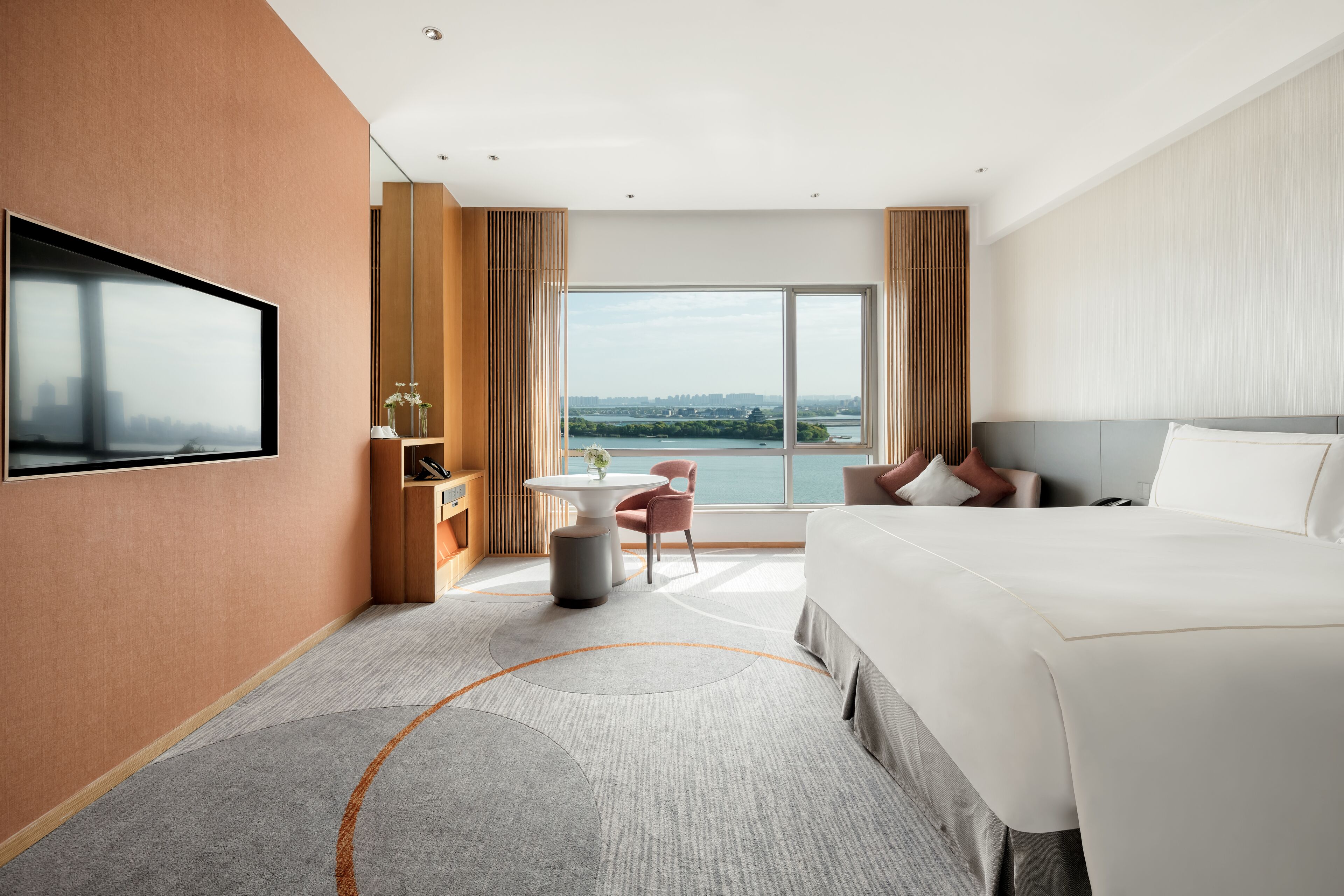A modern hotel room with a large bed, a TV mounted on the wall, a small table with a chair, and a view of the city through large windows.
