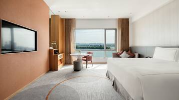 A modern hotel room with a large bed, a TV mounted on the wall, a small table with a chair, and a view of the city through large windows.