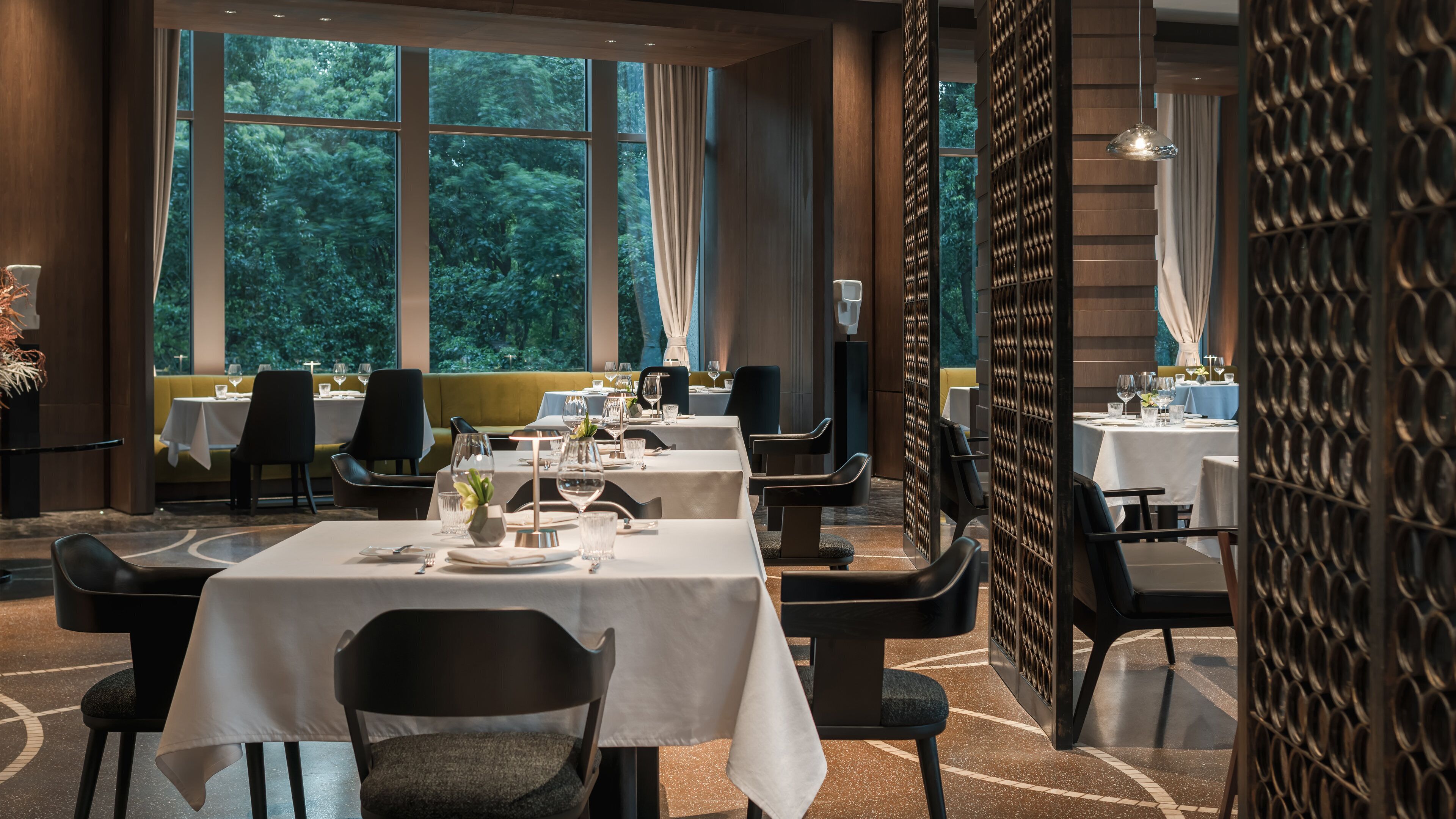 A restaurant with multiple tables set for dining, large windows with a view of trees, and a modern interior design.