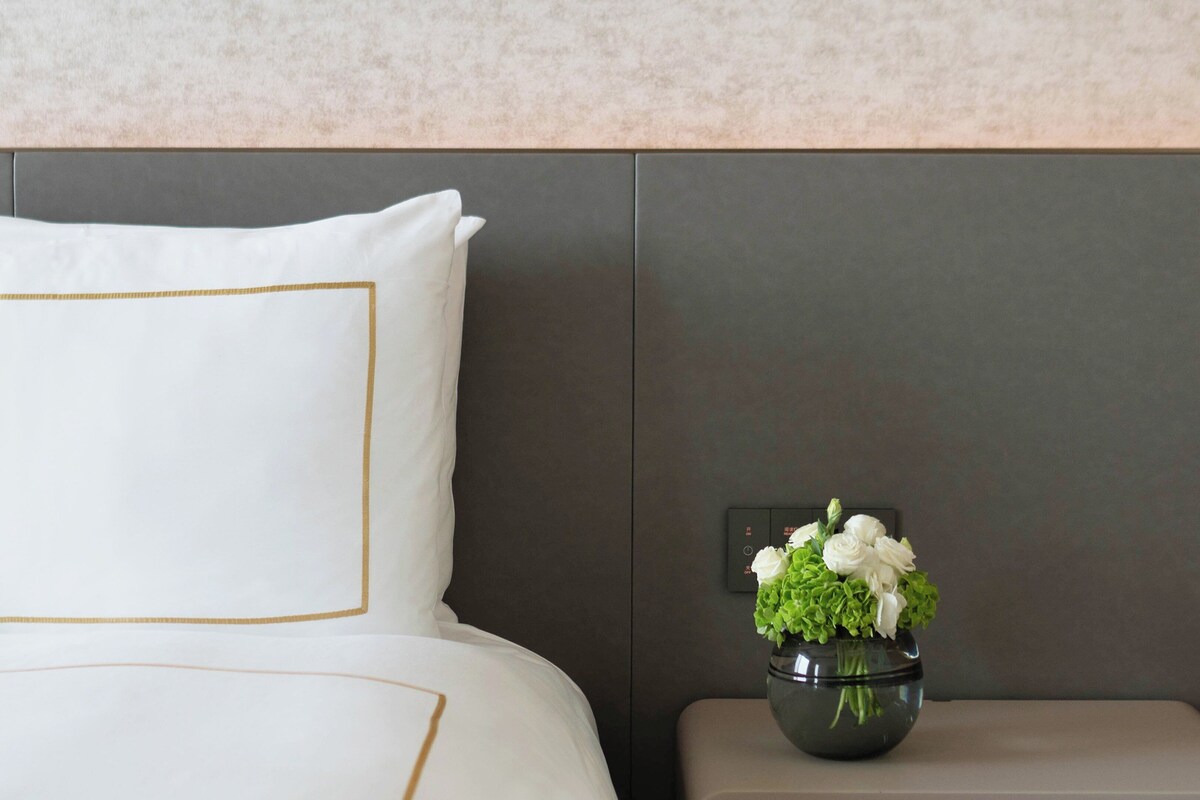 A neatly made bed with a white pillow and a gold trim, a small bedside table with a vase of flowers, and a wall-mounted control panel.