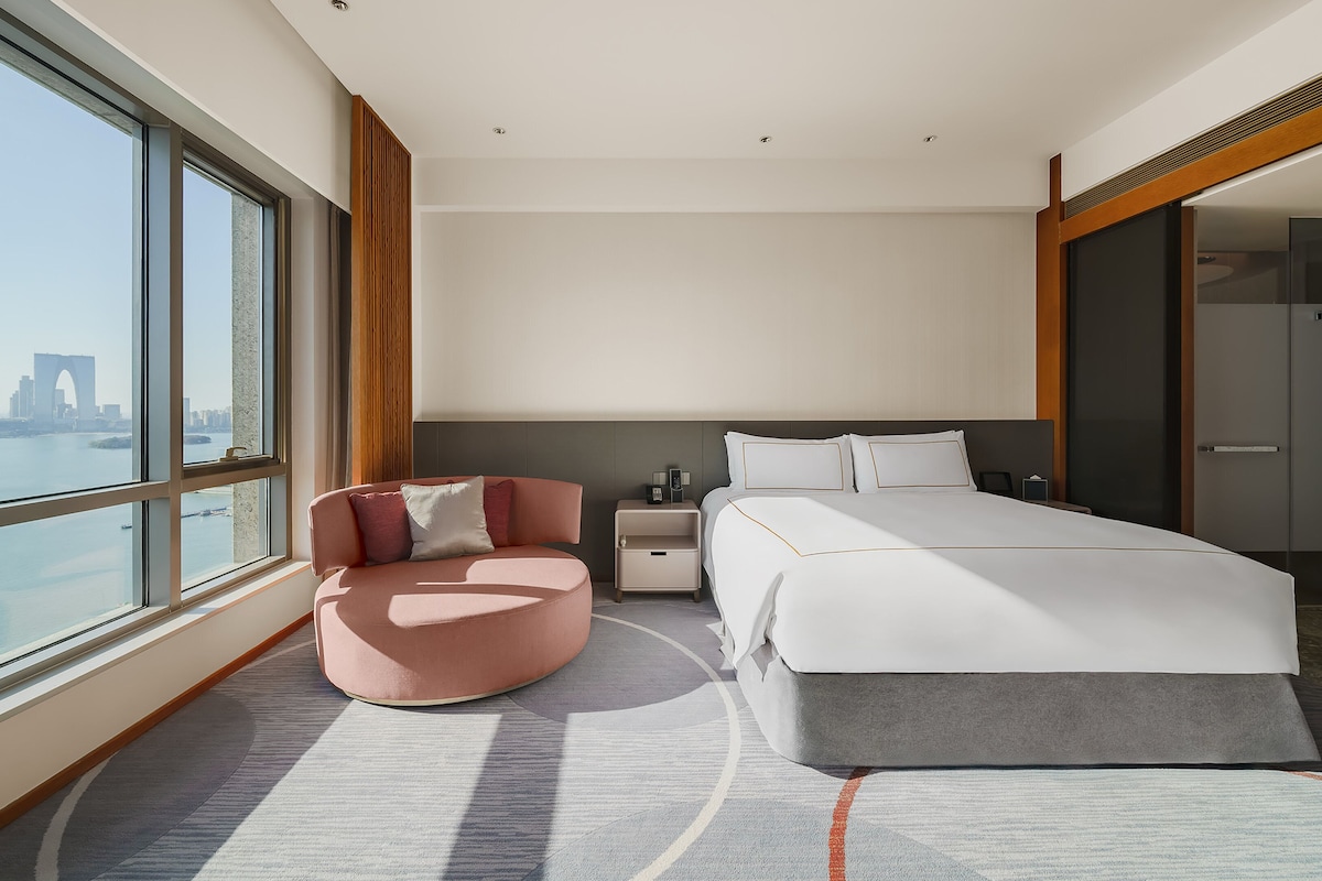A modern hotel room with a large bed, a seating area, and a view of a city skyline.