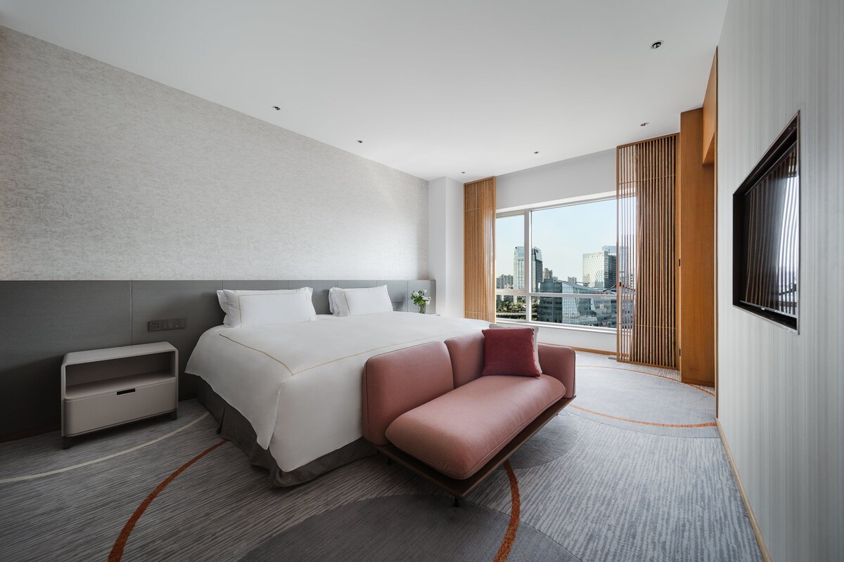 Una habitación de hotel moderna con una cama grande, un sofá, un televisor y vistas a la ciudad.