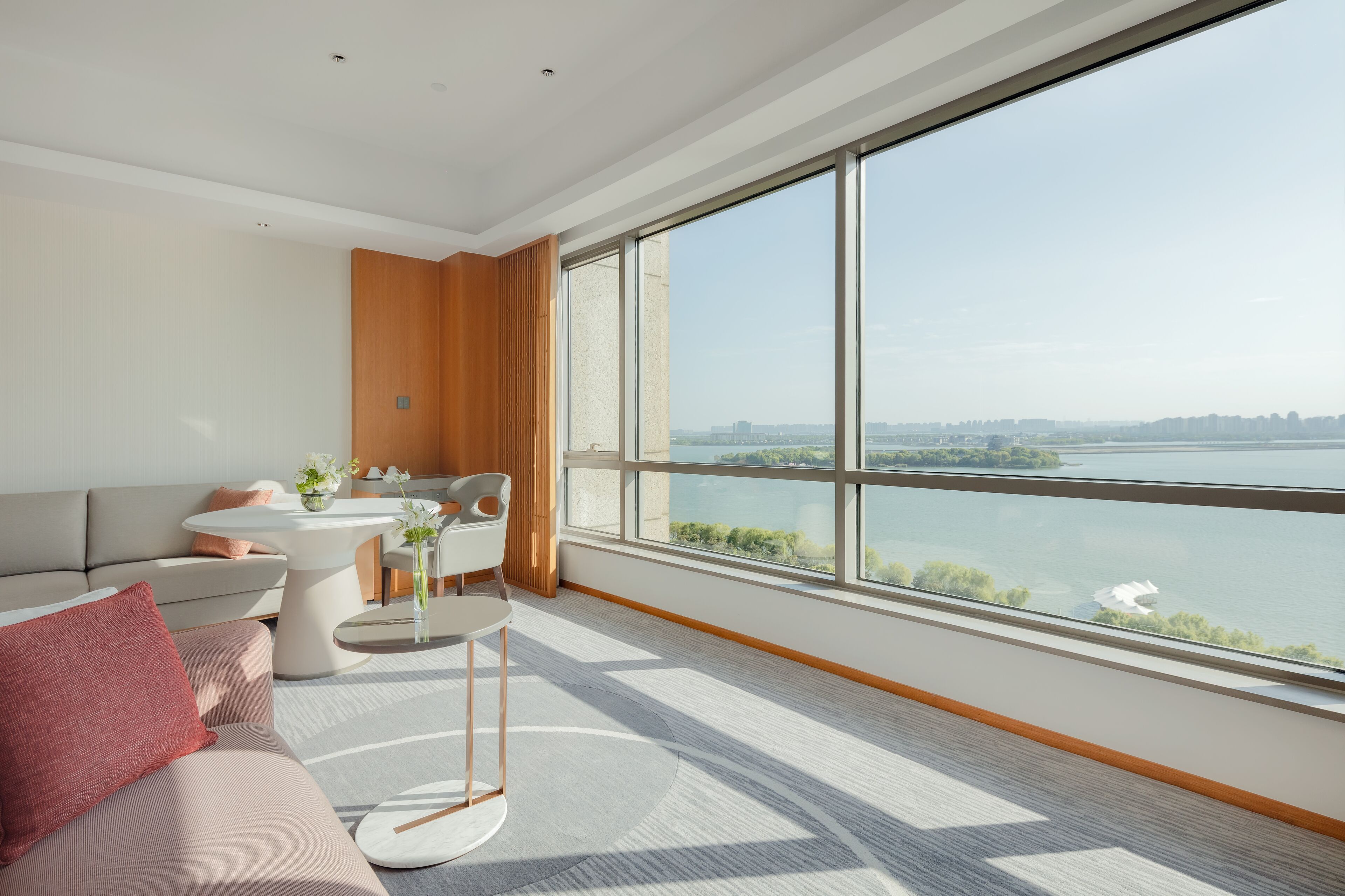 A modern hotel room with a large window offering a city view, a sofa, a small table with a vase of flowers, and a wooden accent wall.