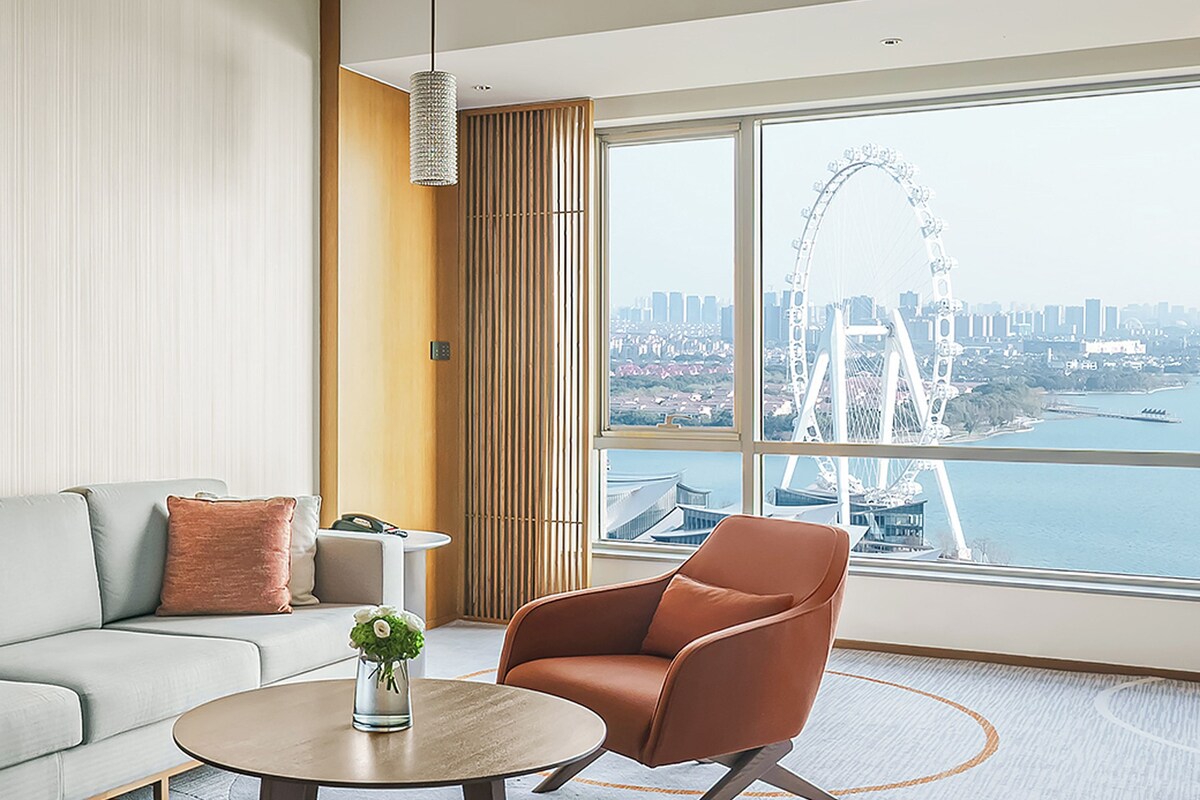 Una habitación de hotel moderna con un gran ventanal, un sofá, una silla y una mesita.