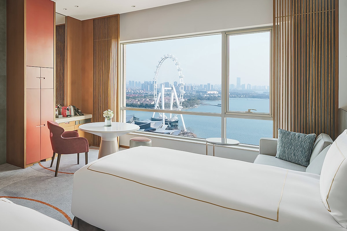 Habitación de hotel con cama, escritorio, silla y un amplio ventanal con vistas a una noria y al horizonte de la ciudad.