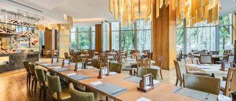 A spacious dining area with long wooden tables and chairs, large windows offering views of the outdoors, and a modern chandelier.