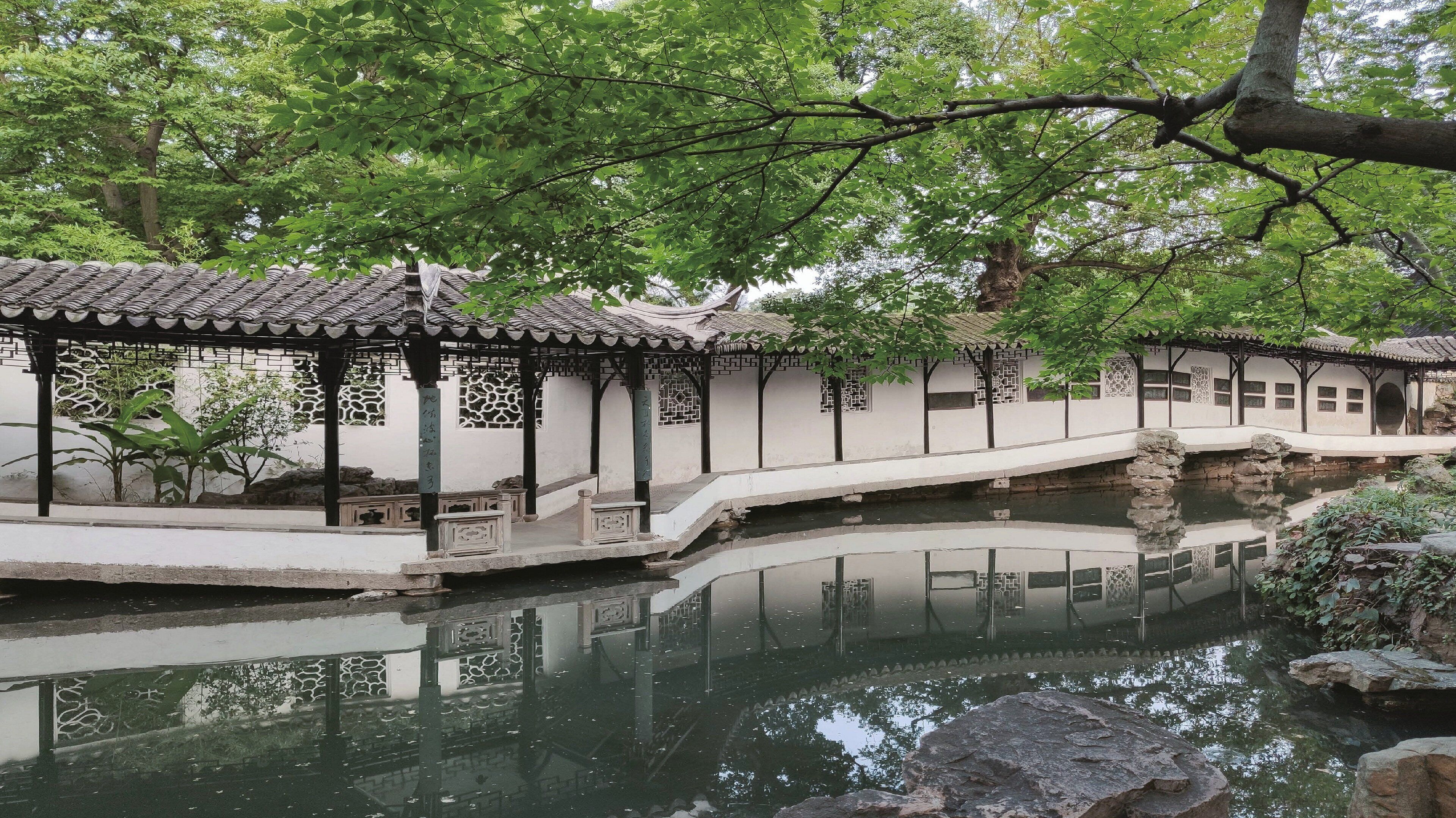A traditional Chinese garden with a stone bridge over a pond, surrounded by lush greenery and classical architecture.