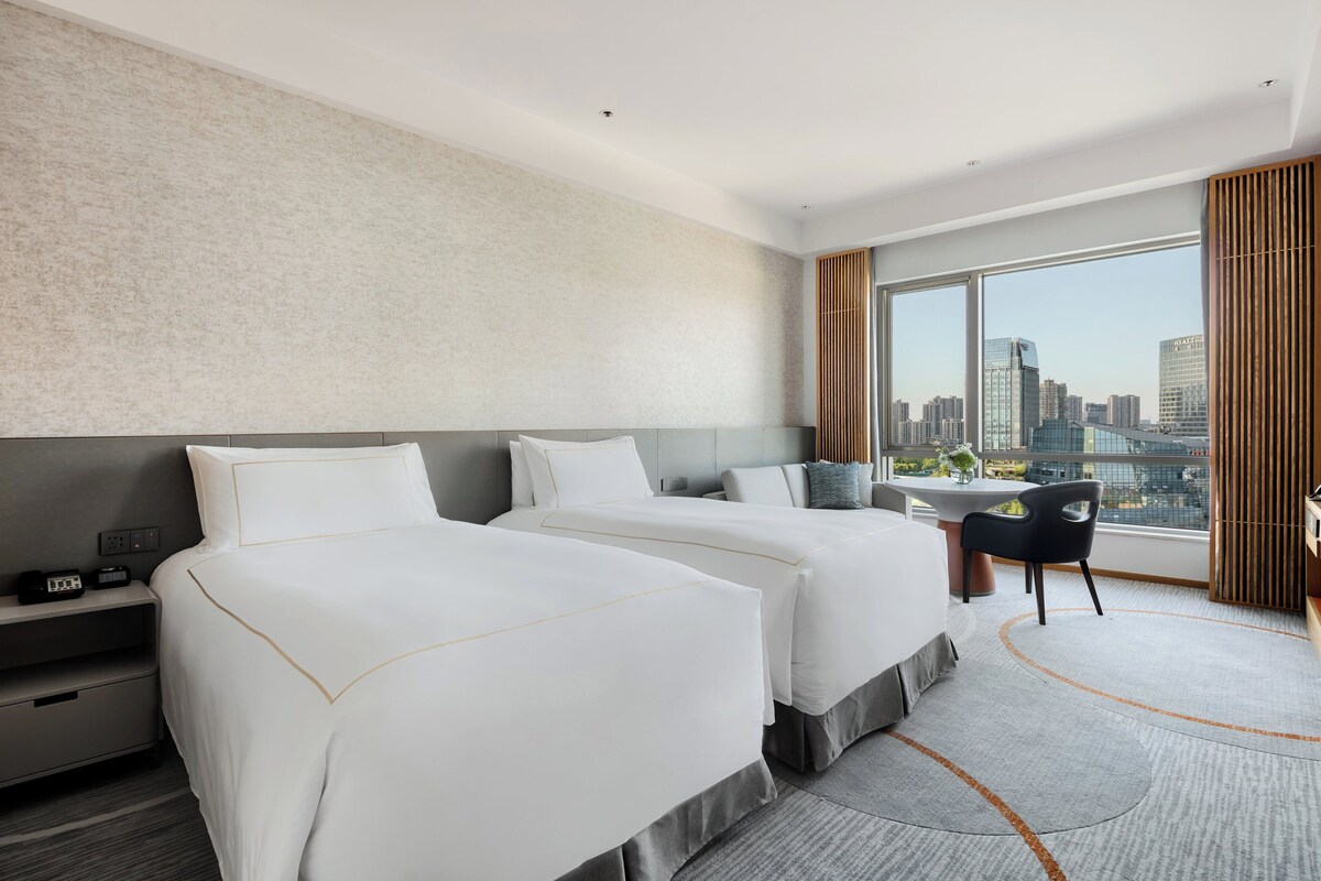 A modern hotel room with two beds, a desk, and a large window offering a city view.