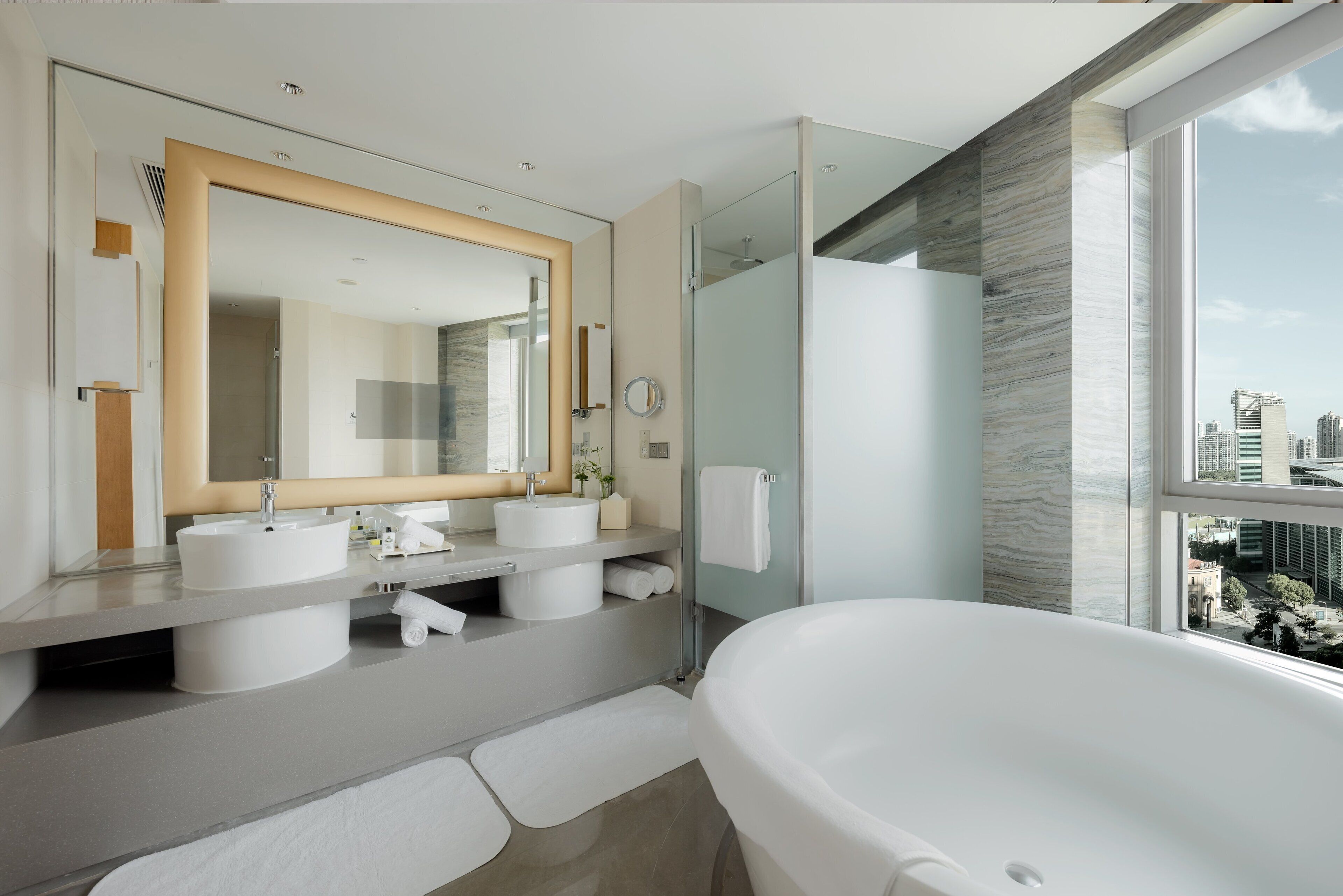 A modern bathroom with a large bathtub, two sinks, a glass shower enclosure, and a city view.
