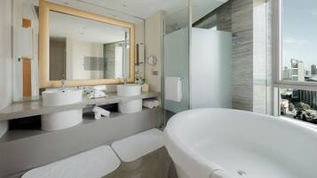A modern bathroom with a large bathtub, two sinks, a glass shower enclosure, and a city view.