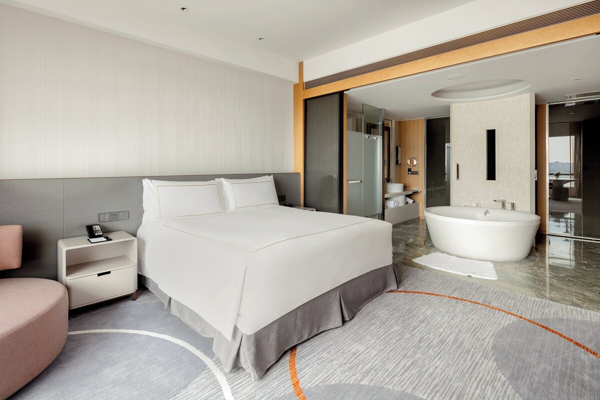 Une chambre d’hôtel moderne avec un grand lit, une table de chevet avec un téléphone, un petit coin salon et une salle de bain dotée d’une baignoire et d’un grand miroir.