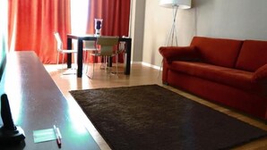 Una sala de estar con un sofá rojo, una mesa de comedor con sillas, una lámpara y un tapete grande.