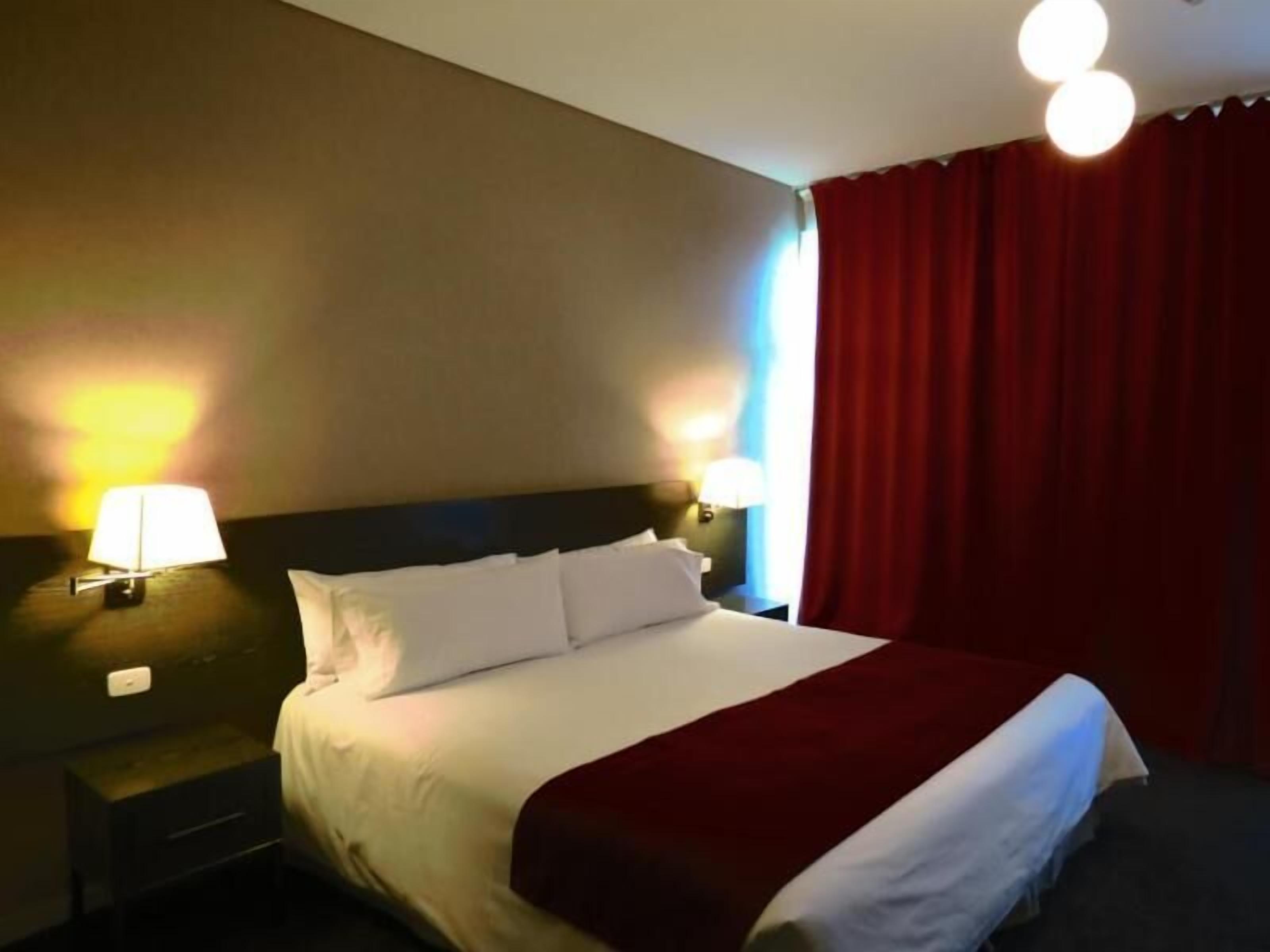 A hotel room with a bed, two bedside lamps, a nightstand, and a red curtain.
