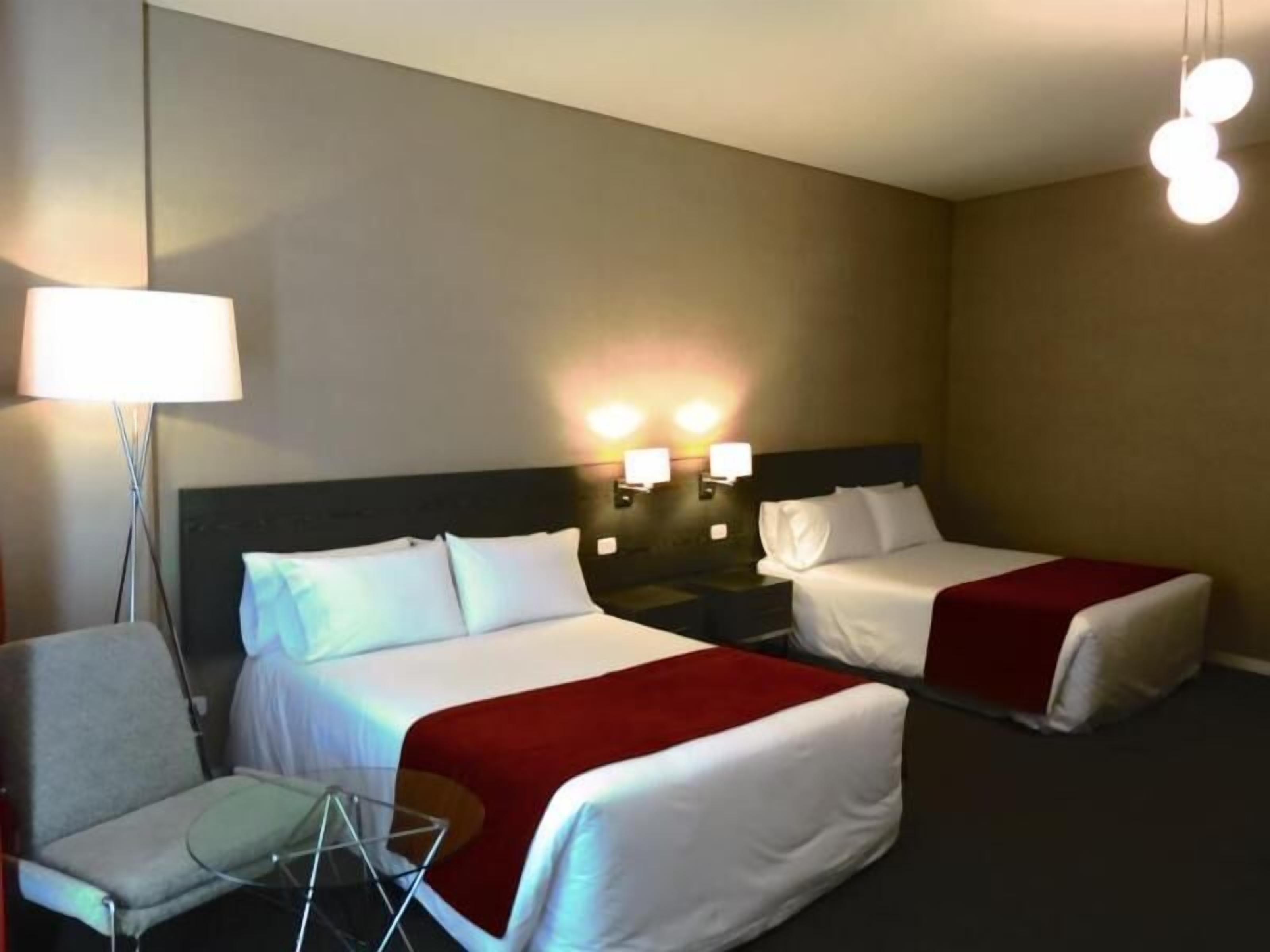 A hotel room with two beds, a chair, a lamp, and wall-mounted lights.