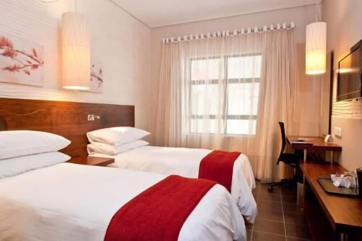 A hotel room with two beds, a desk, a chair, and a large window with curtains.