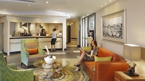 A hotel lobby with a reception desk, a receptionist, a waiting area with a sofa and chairs, and a coffee table with a bowl and plant. - Peermont Metcourt Hotel at Umfolozi (Empangeni)