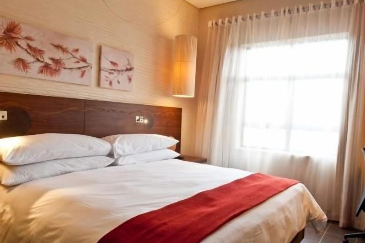 A hotel room with a bed, a headboard, a wall-mounted light, and a window with curtains.