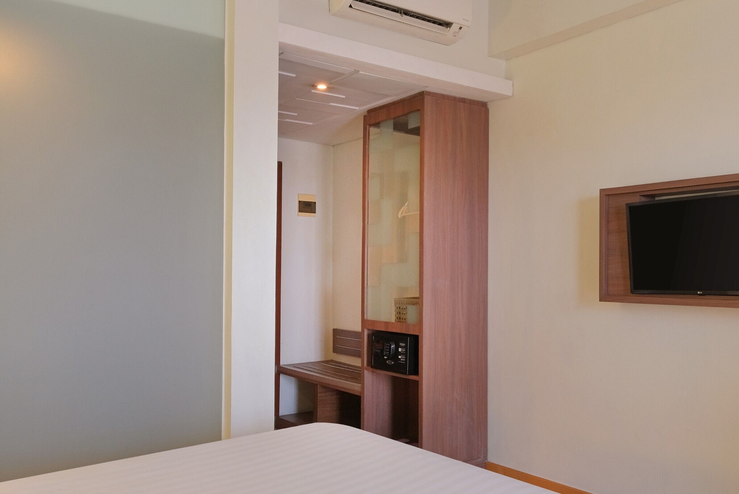 A hotel room with a bed, a wooden wardrobe, a television mounted on the wall, and a bench.