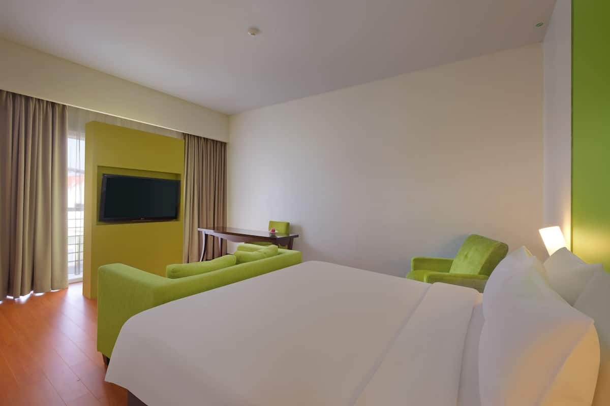 Ein Hotelzimmer mit einem großen Bett, einem grünen Sessel, einem kleinen Tisch und einem an der Wand befestigten Fernseher.