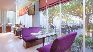 A modern indoor dining area with purple upholstered seating, a long table with cutlery, and large windows overlooking an outdoor seating area.
