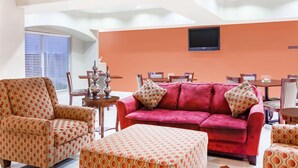 Una sala de estar con un sofá rojo, sillones con estampado y un otoman, un comedor con mesa y sillas, y un televisor de pantalla plana montado en una pared naranja.