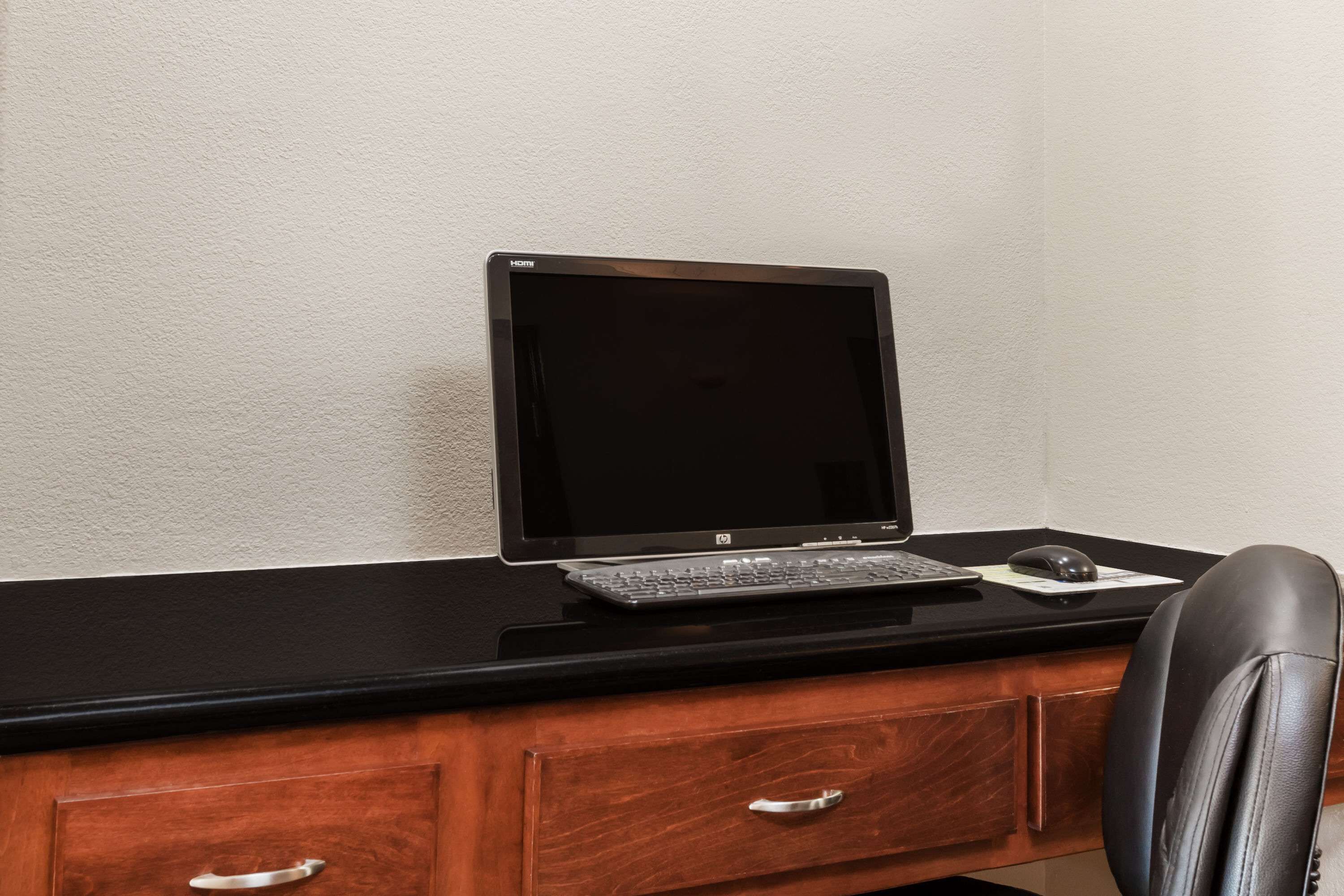 Un monitor HP nero su una scrivania in legno, con una tastiera e un mouse neri, una poltrona in pelle e un mobiletto portaoggetti in legno.
