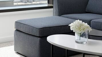 A modern living room with a grey sofa and ottoman, a clear glass vase with white flowers on a white table, and a city view through large windows with grey curtains.