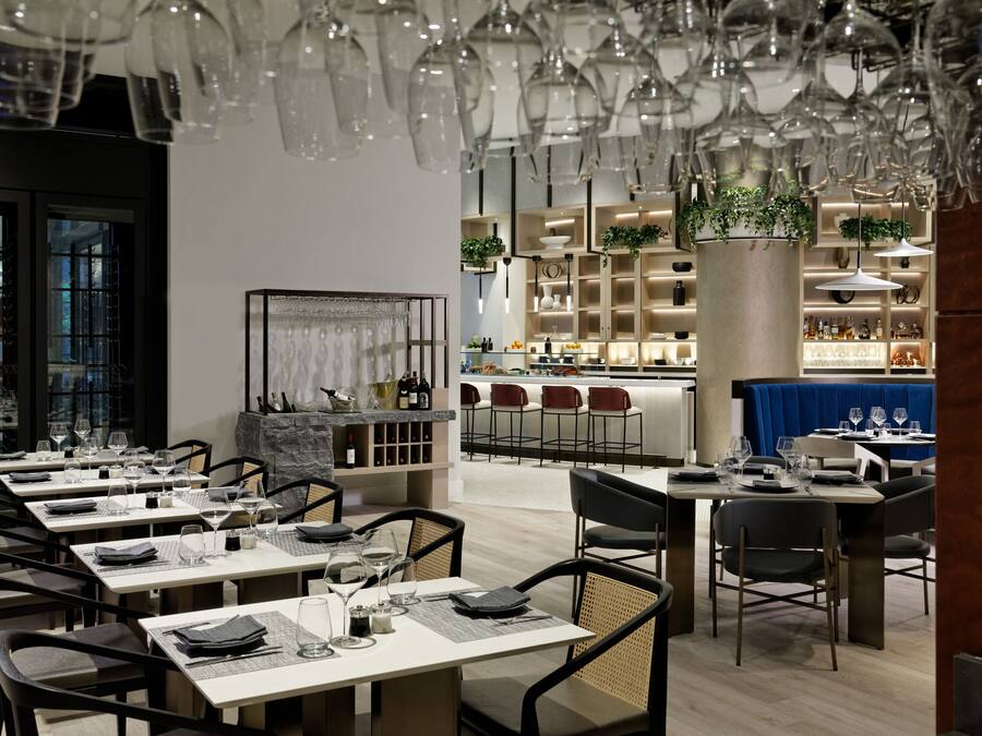 A modern restaurant with multiple dining tables and chairs, a bar area, and a decorative ceiling with hanging glassware.