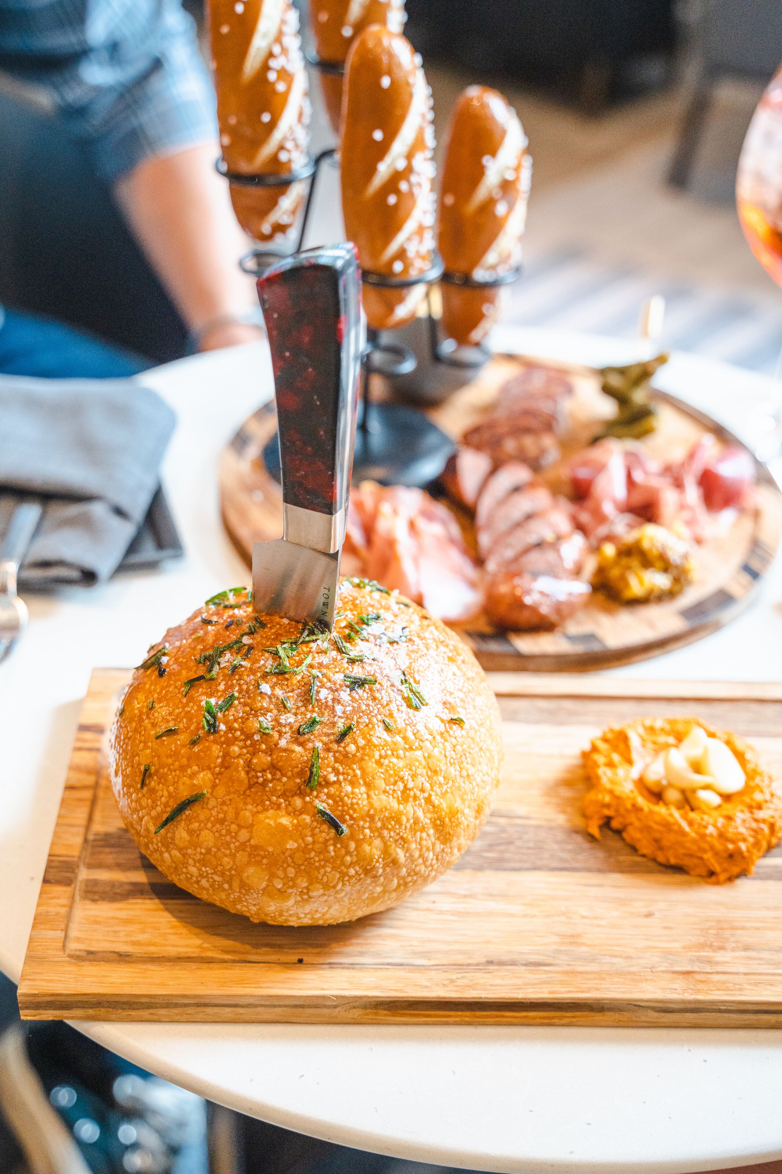 A round, sesame seed-covered bread on a wooden board, a knife cutting through it, and a plate with sliced meats and pretzels in the background.
