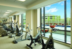 A gym with various exercise machines, large windows, and a view of a building and trees outside.