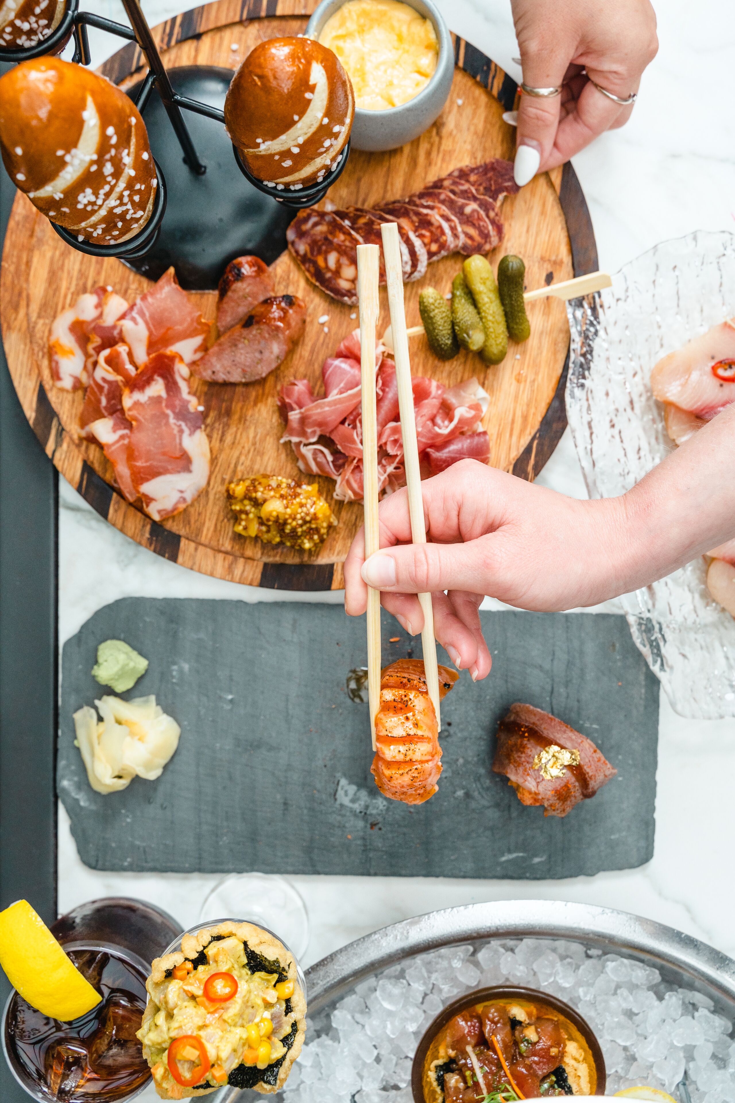 A variety of food items on a wooden board, including meats, cheeses, and pickles, with a person using chopsticks to pick up a skewer.