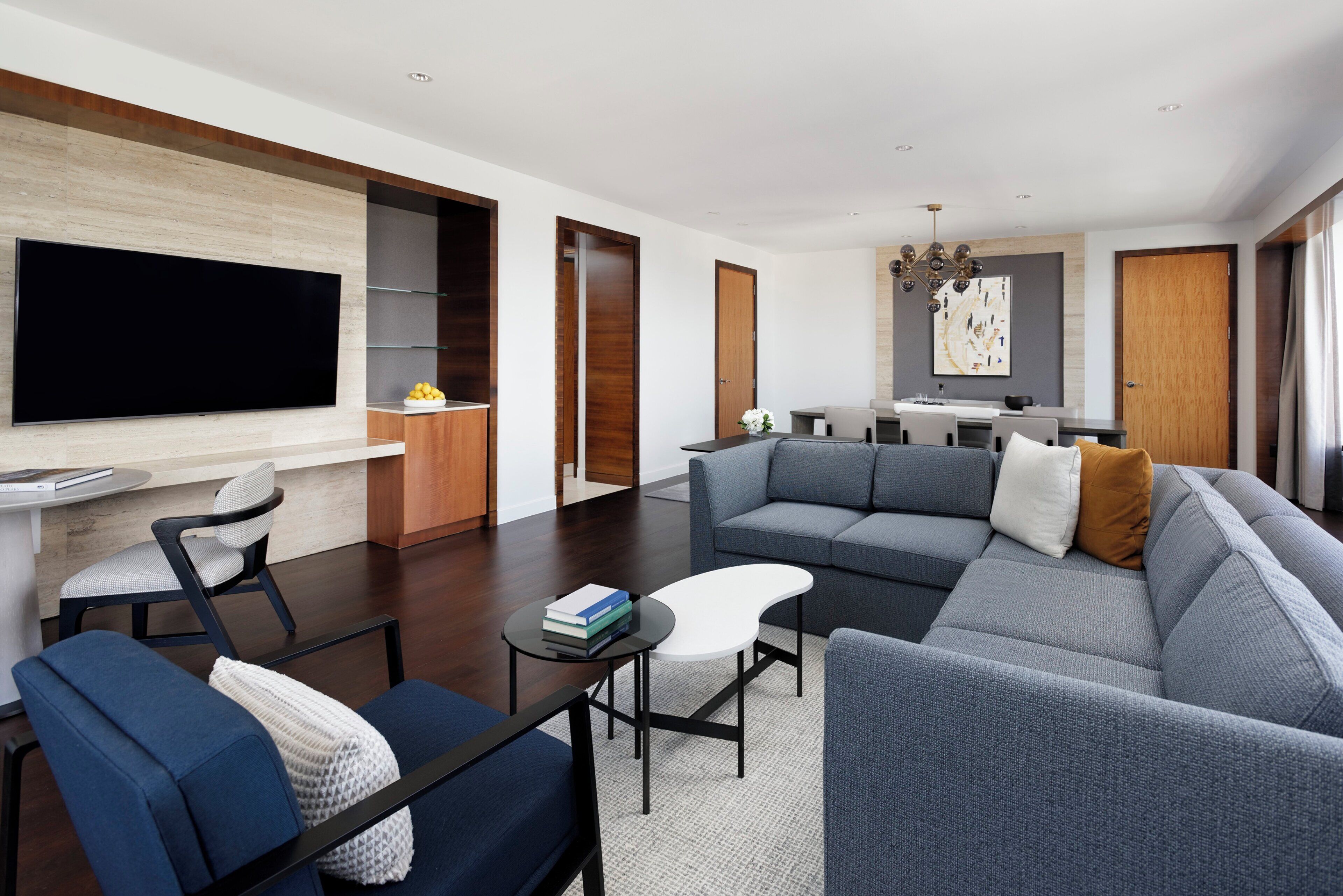 A modern living room with a large flat-screen TV, a grey sectional sofa, a blue armchair, and a small coffee table.