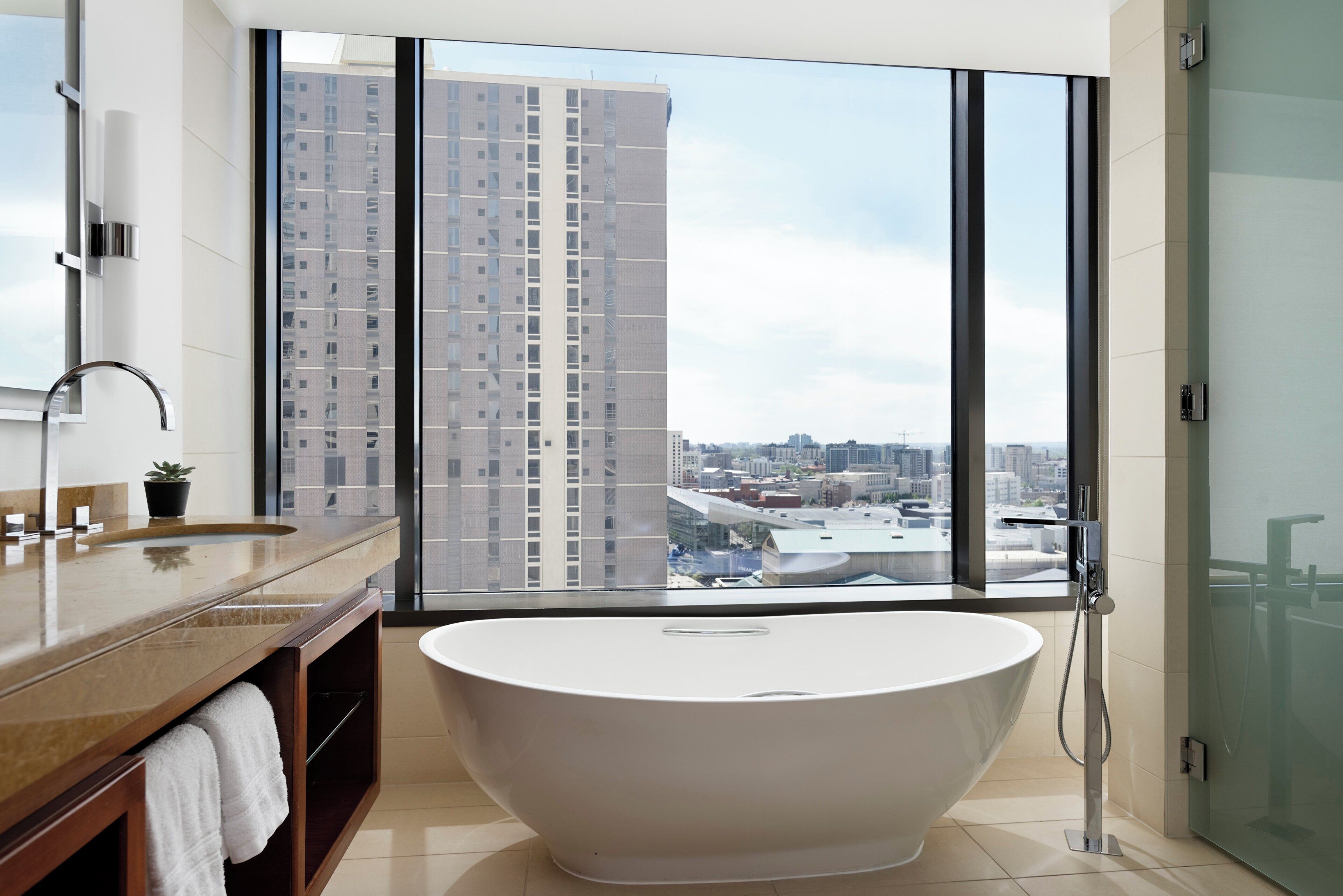A modern bathroom with a freestanding bathtub, a large window with a city view, and a vanity with a mirror and sink.