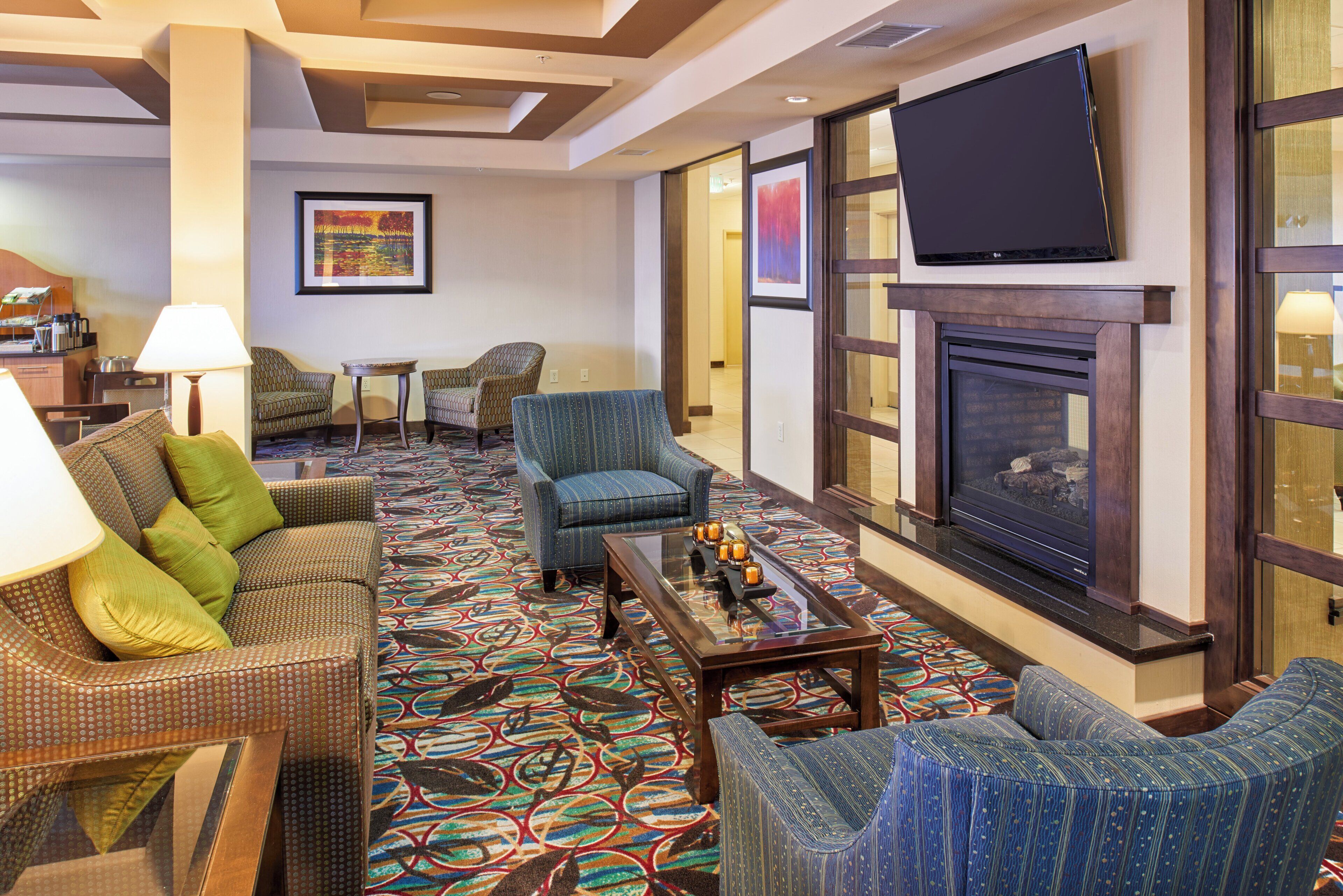 A hotel lounge with a patterned carpet, wicker furniture, a flat-screen TV, and a fireplace.