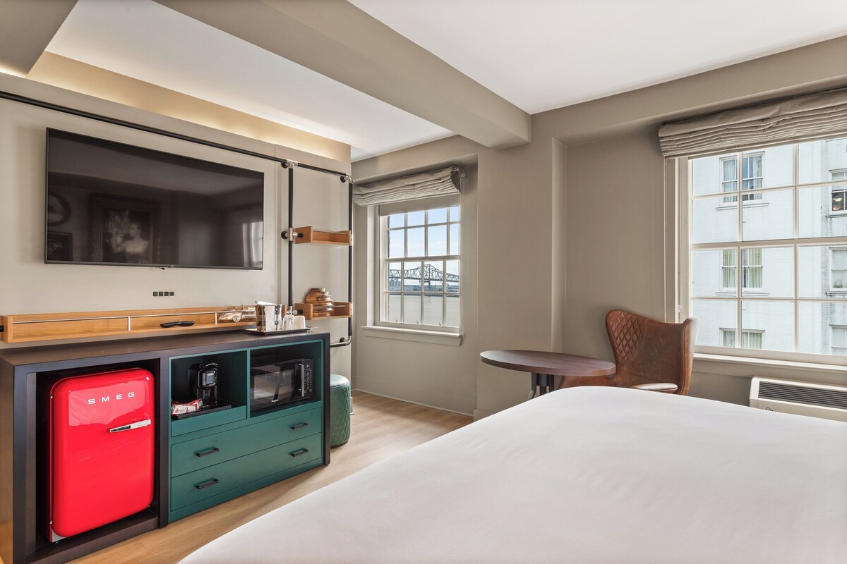 A hotel room with a bed, a small kitchenette with a red fridge, a TV mounted on the wall, and a window with a view of buildings.