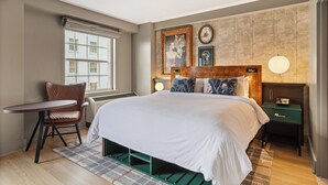 A modern bedroom with a large bed, a desk, and a chair. There is a window with blinds, a wall clock, and framed pictures.