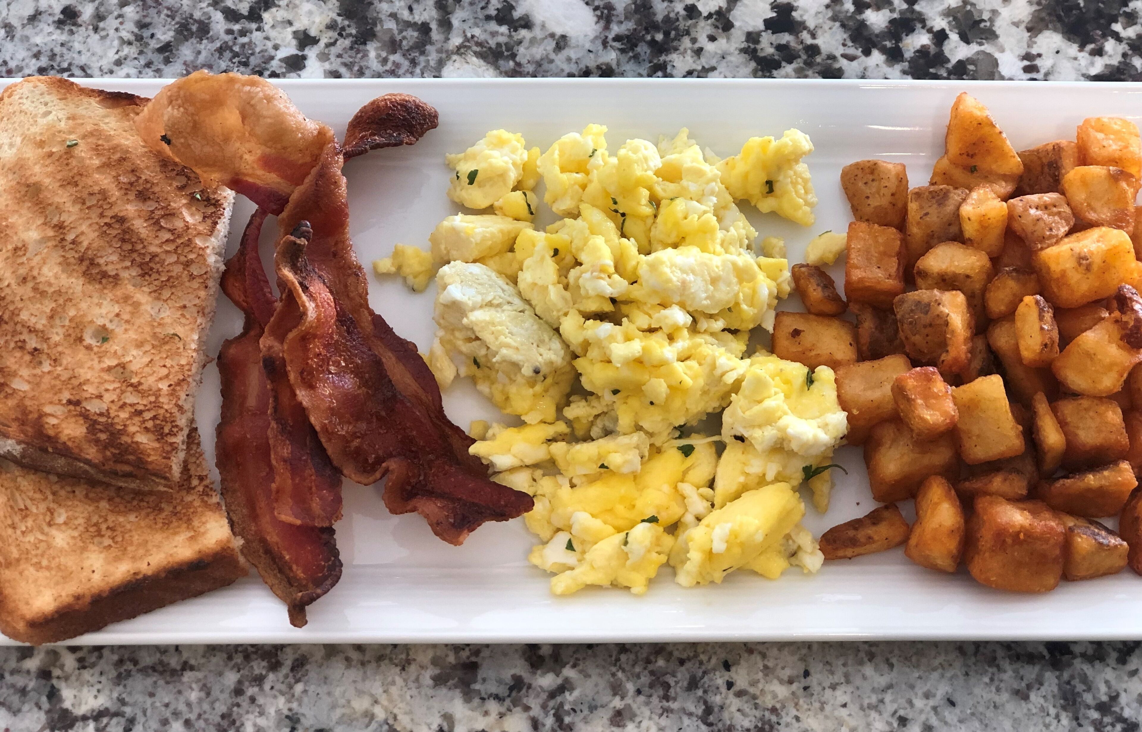 Prato de café da manhã com torradas, bacon, ovos mexidos e batatas assadas.
