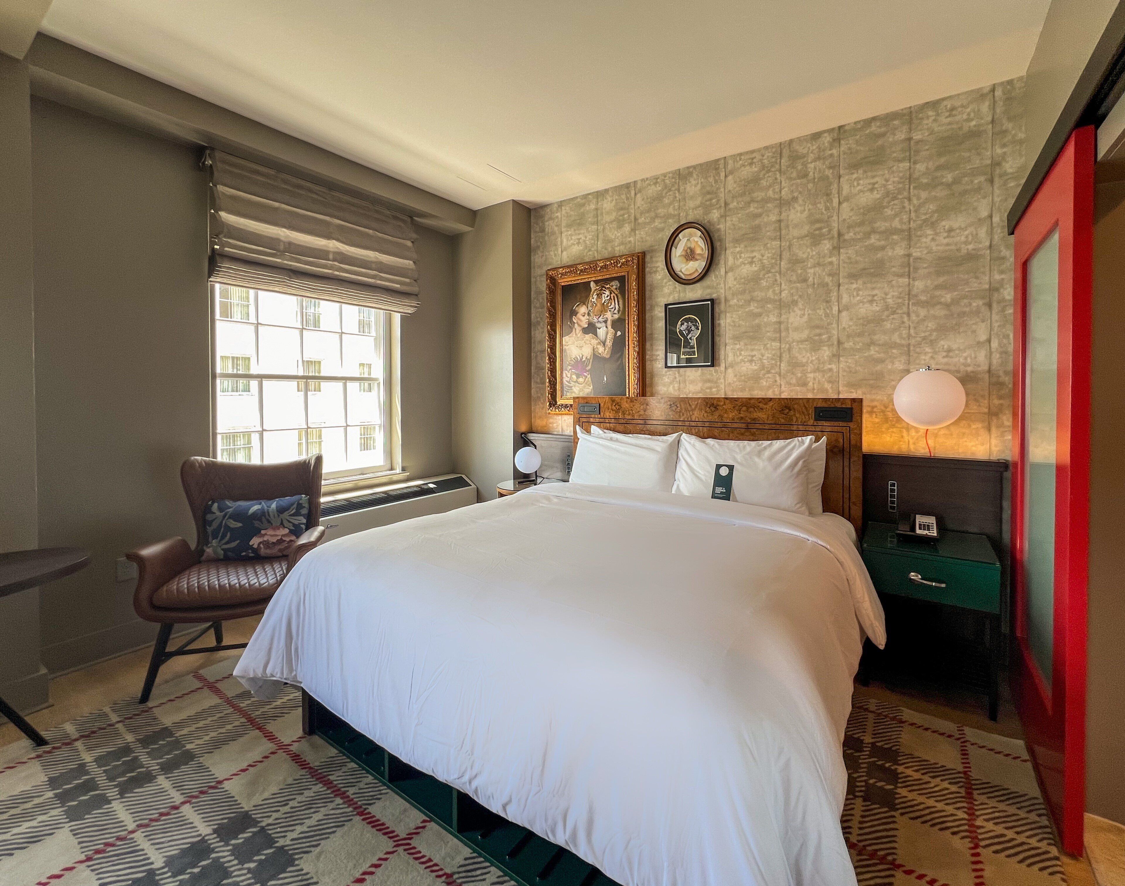 A hotel room with a large bed, a chair, a nightstand, a wall clock, and a window with blinds.