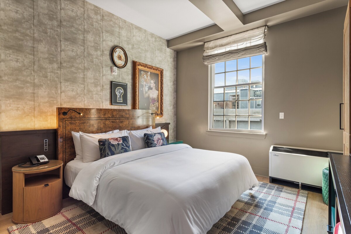 A neatly arranged hotel room with a large bed, a bedside table with a lamp, a window with a view of buildings, and a wall-mounted heater.