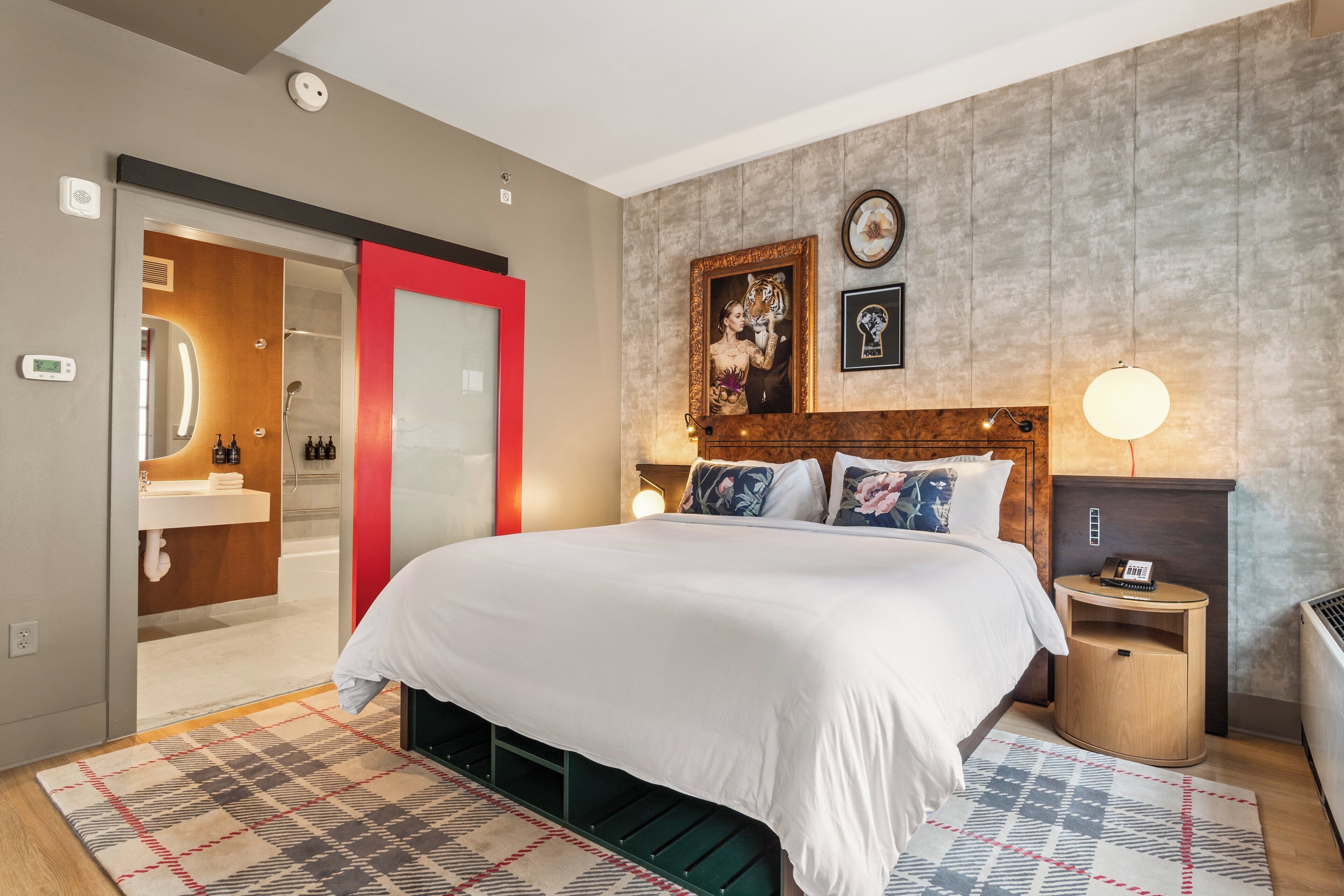Una habitación de hotel moderna con una cama grande, cabecera de madera, mesita de noche con lámpara, espejo con marco rojo y un baño visible a través de una puerta abierta.