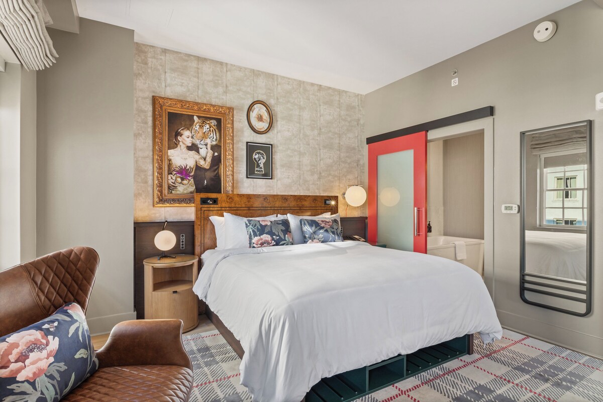 A modern bedroom with a large bed, a wooden headboard, a nightstand with a lamp, a red wardrobe, and a mirror.