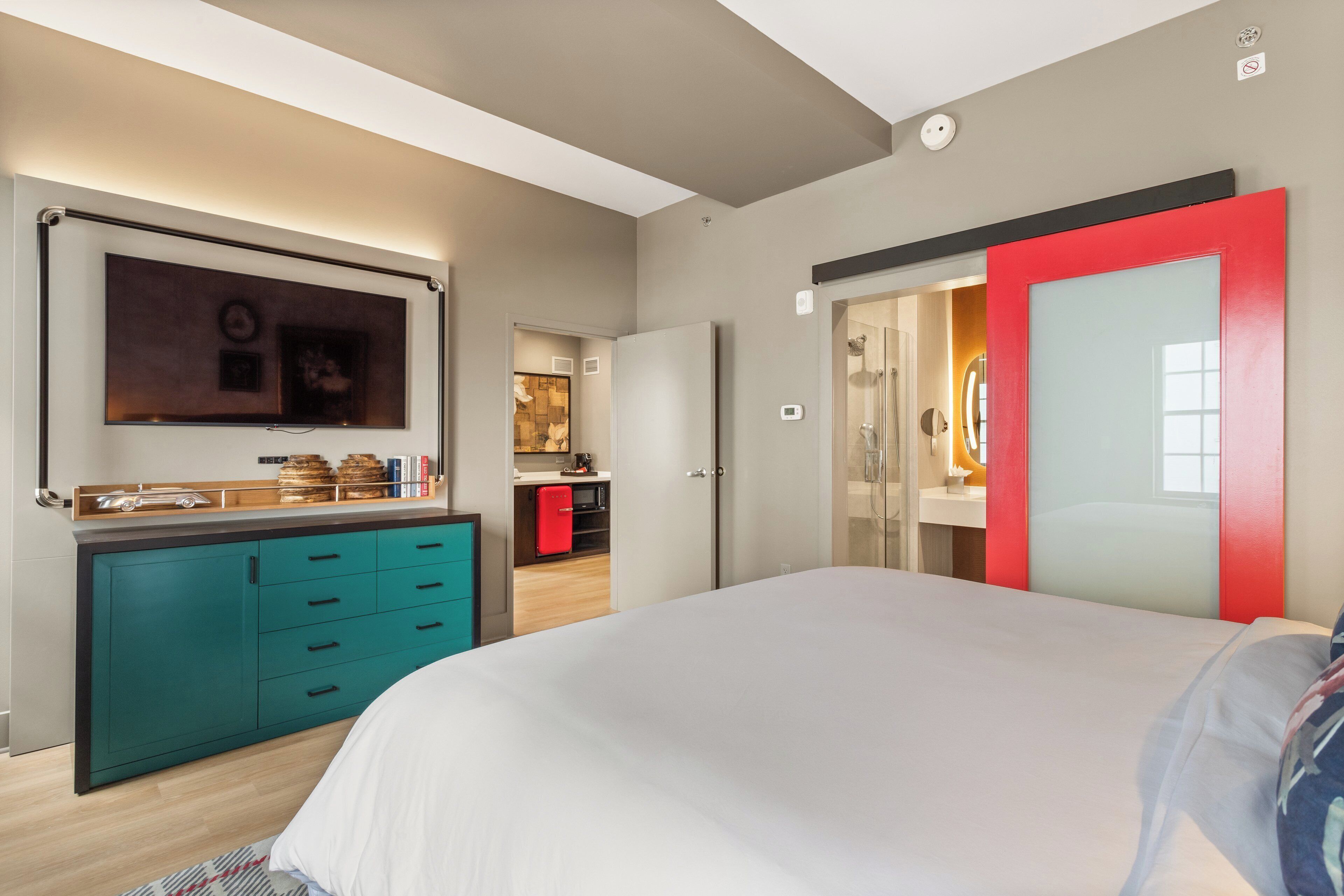 A modern bedroom with a large bed, a teal dresser, a red-framed mirror, and a small kitchenette in the background.