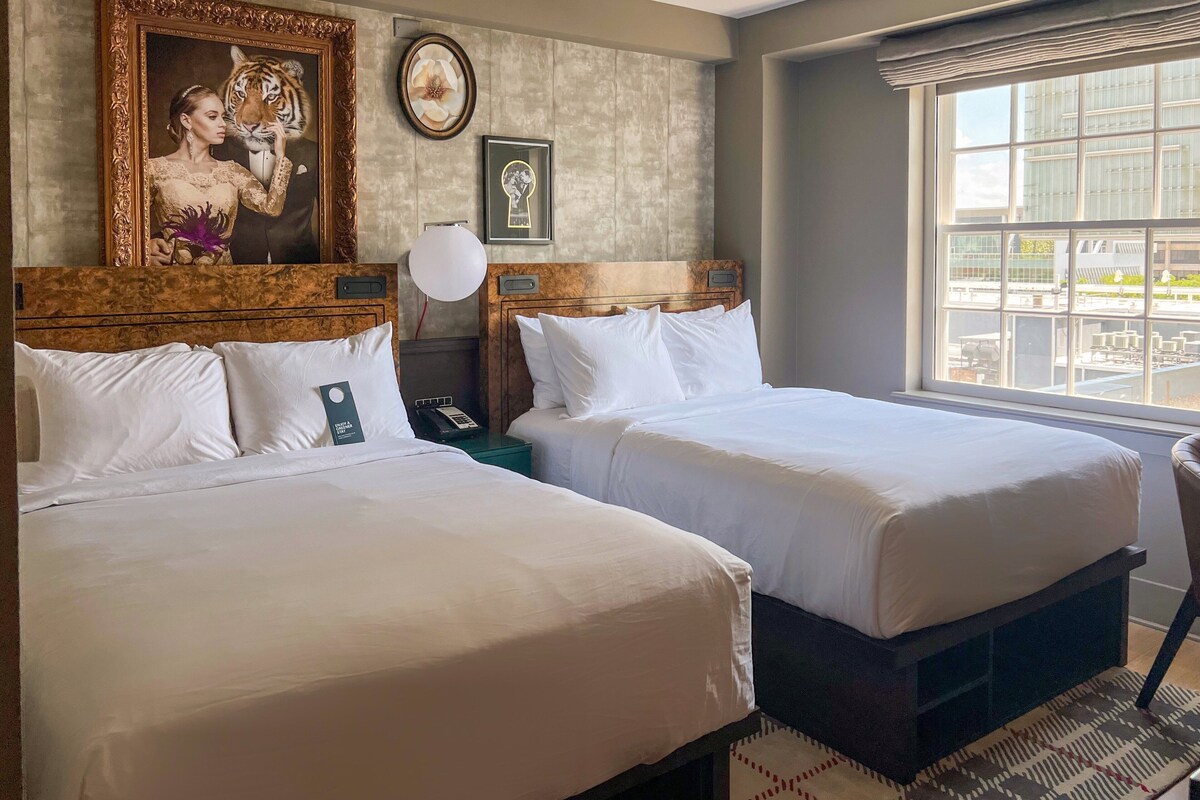 A hotel room with two beds, a large window with a city view, a framed picture of a tiger, and a modern lamp.