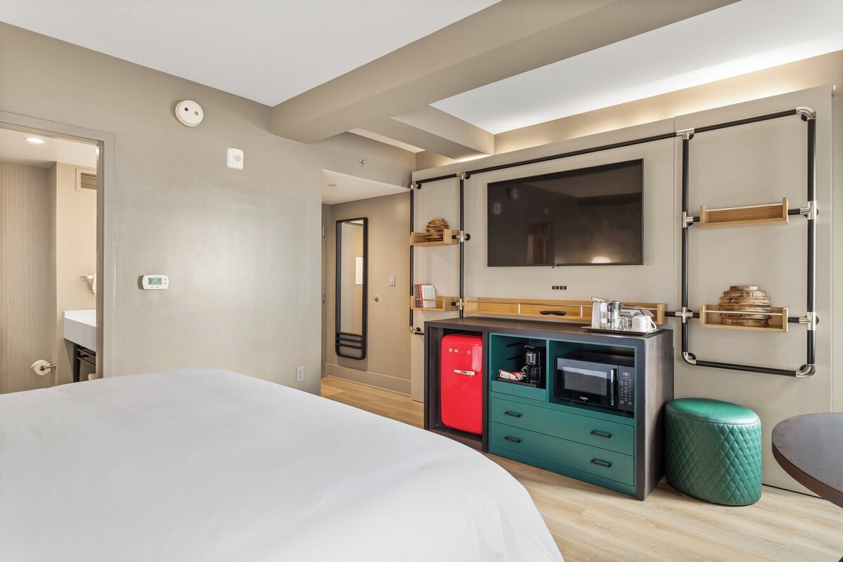 A modern hotel room with a large bed, a kitchenette with a red fridge, a microwave, and a small green ottoman.