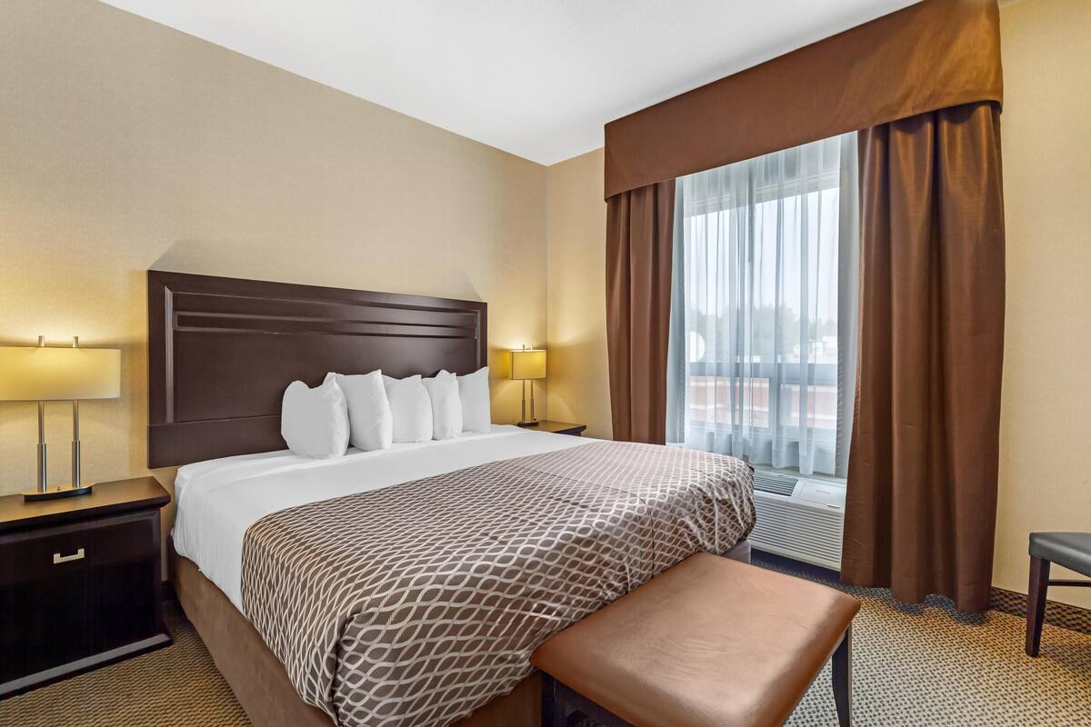 A hotel room with a large bed, two bedside tables with lamps, a window with curtains, and a small bench.
