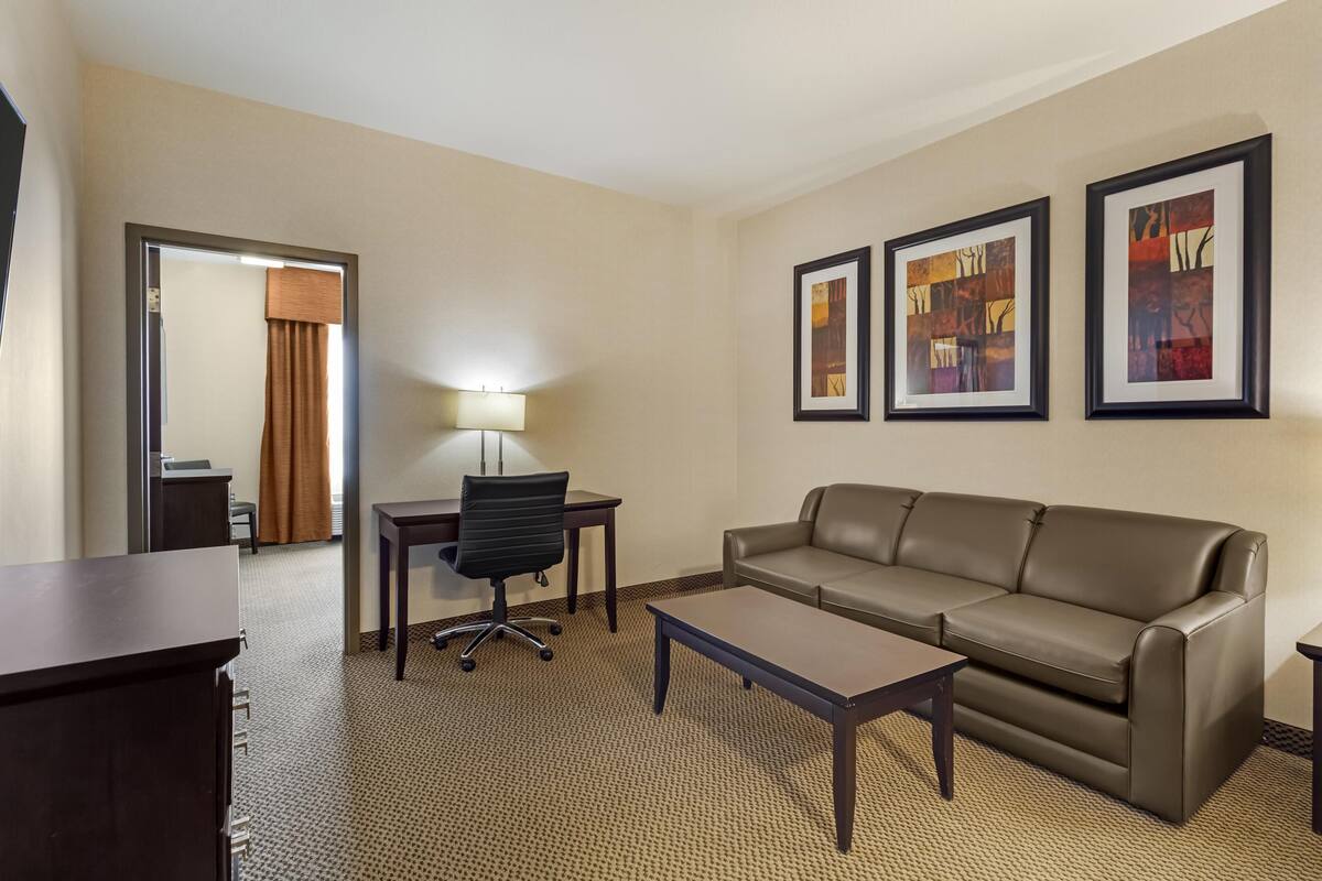 A hotel room with a leather sofa, a desk with a chair, a lamp, and three framed artworks on the wall.