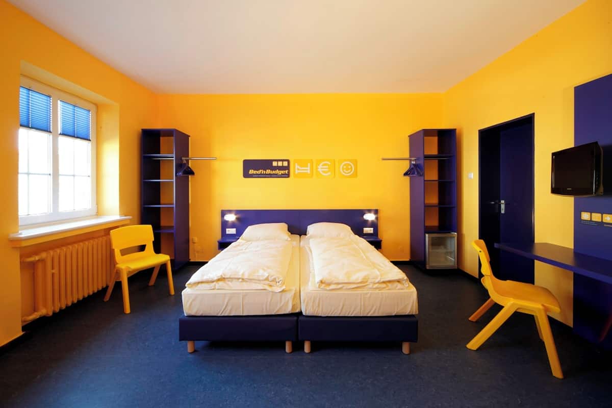 A room with two beds, a window with blinds, a radiator, a desk, a chair, and a television on the wall.