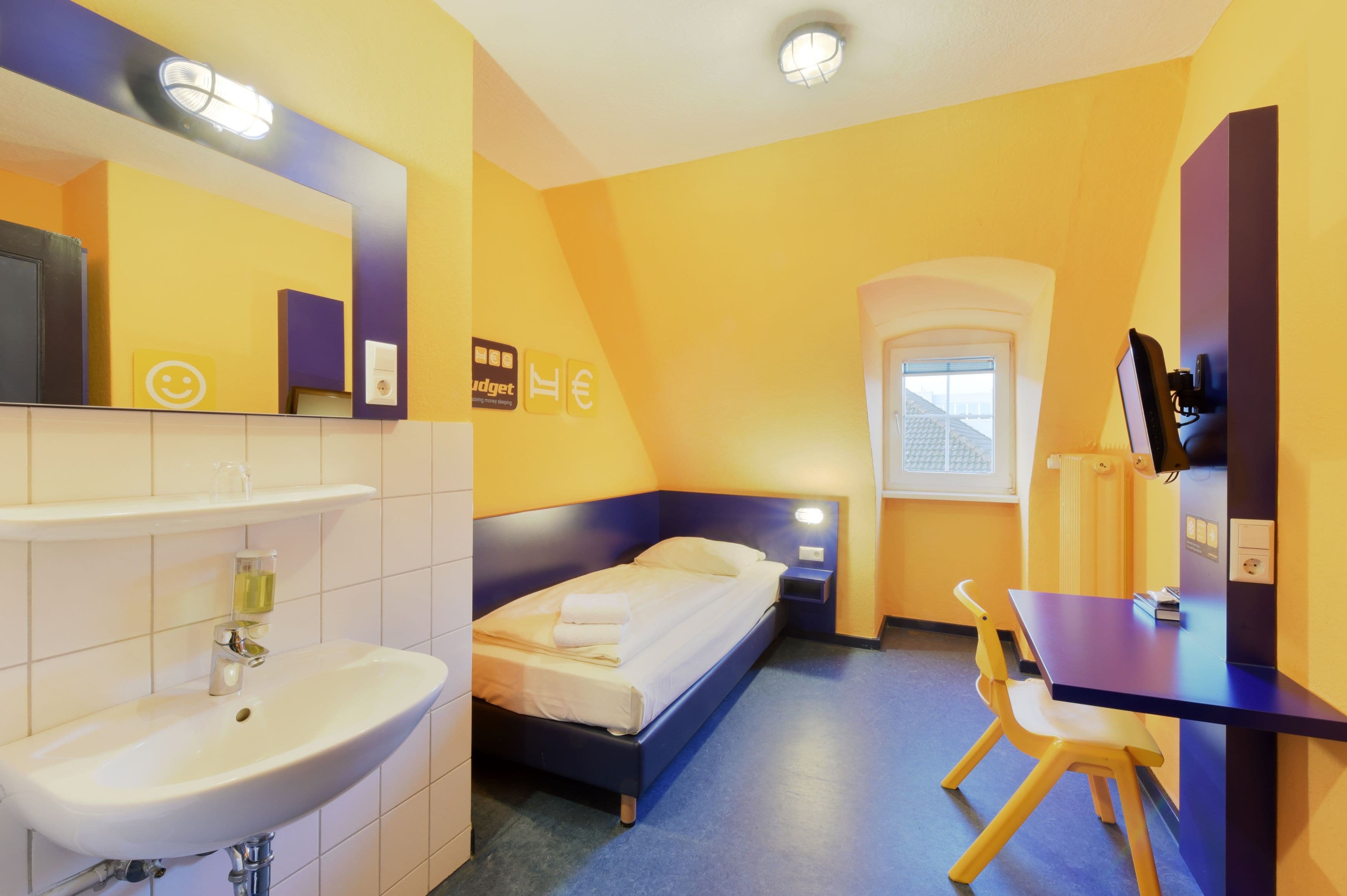 Habitación de hotel con cama, escritorio, silla, lavamanos y ventana.